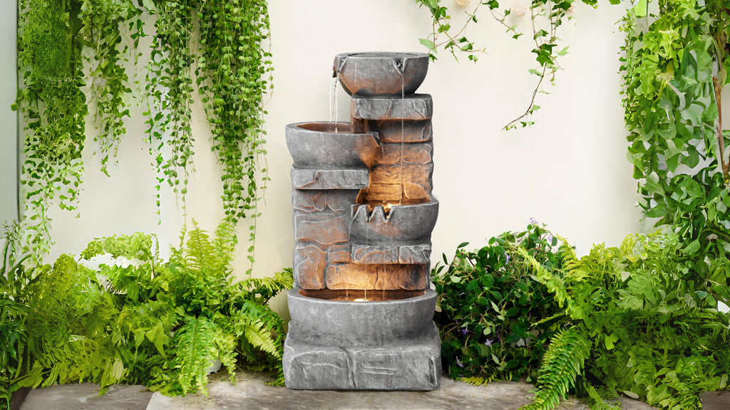 A three-tier stone water fountain in front of a white wall surrounded by green plants and leaves